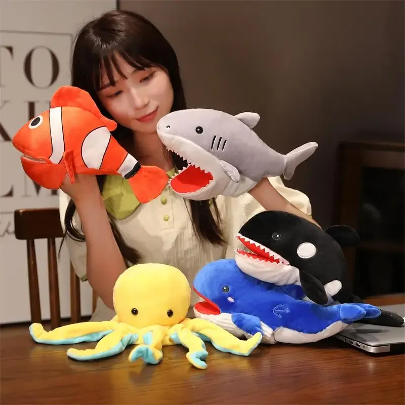 Person holding and displaying a collection of marine animal hand puppets, including a clownfish, shark, orca whale, octopus, and blue fish.